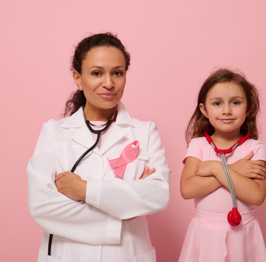 Gorgeous woman doctor in white medical gown stands next to a cute little girl in pink, both with crosses arms on chest and wearing a pink ribbon, symbol of Breast Cancer Awareness Day. Medical concept